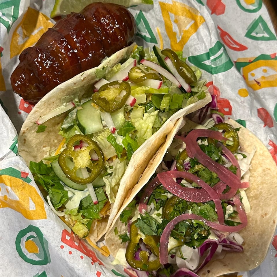 Two Krazy Taco 937 tacos with various toppings and a large glazed sausage on patterned paper.