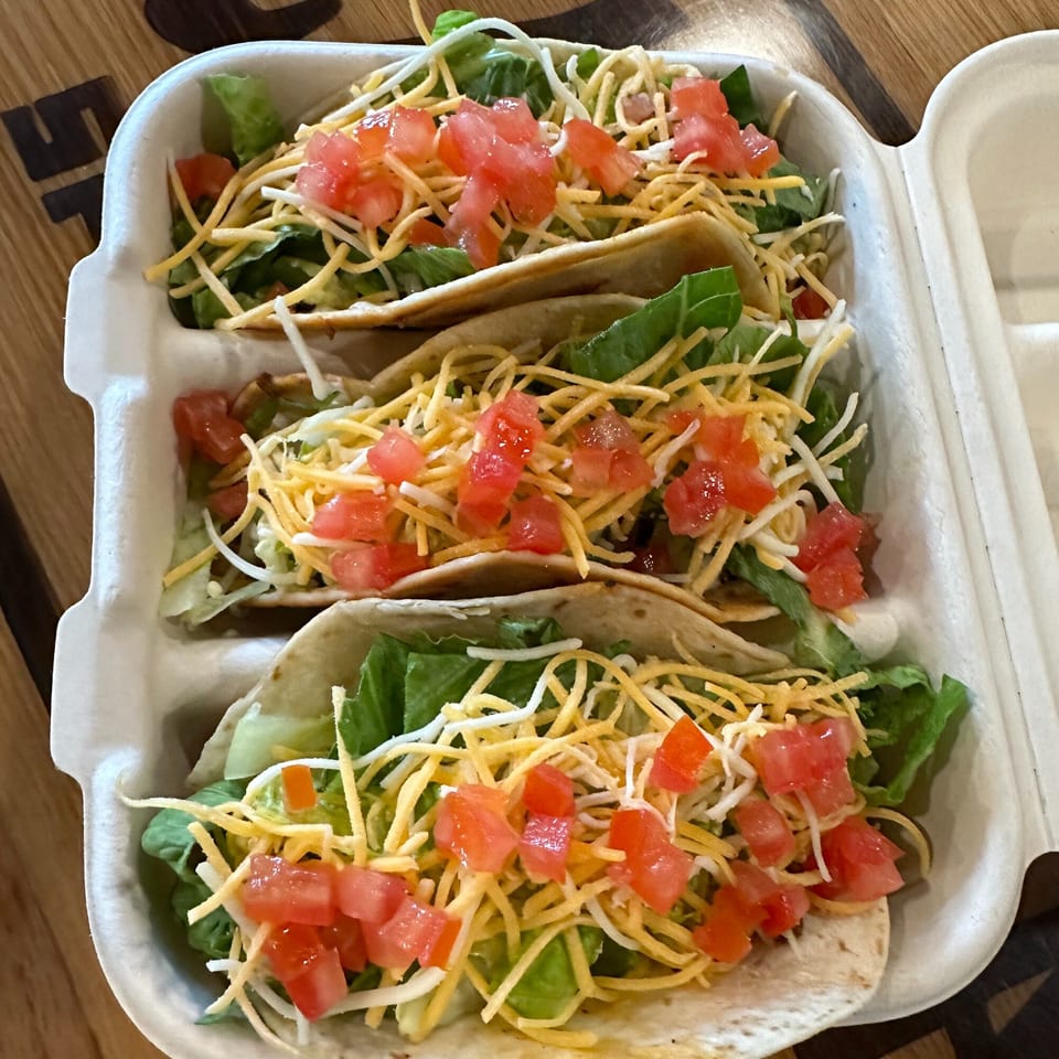 Three Krazy Taco 937 tacos in a white container, topped with lettuce, shredded cheese, and diced tomatoes.