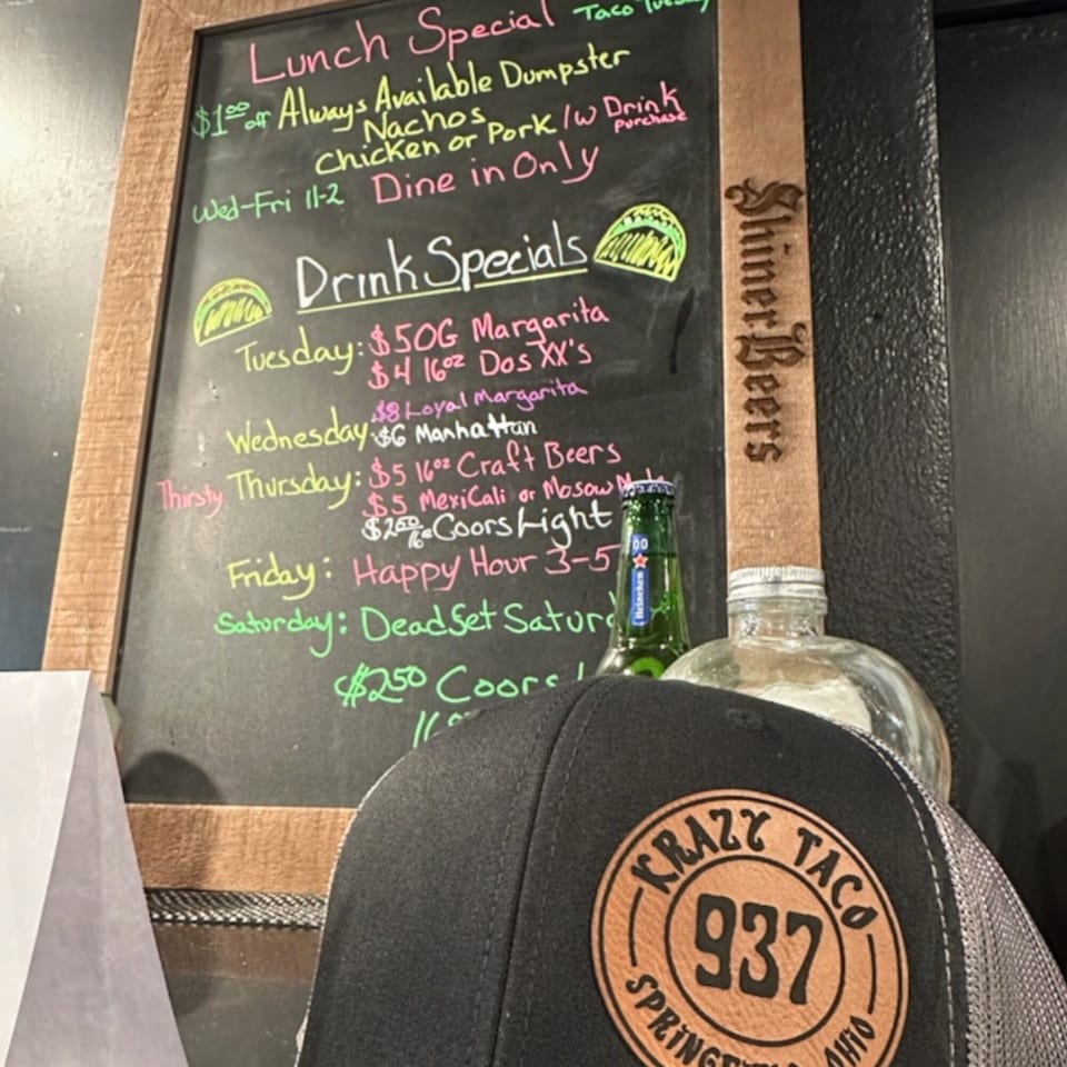 Krazy Taco 937 hat, beer bottle, and a chalkboard with lunch and drink specials.