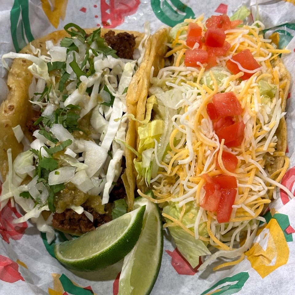 Two Krazy Taco 937 tacos, one with green salsa and the other with cheese and tomatoes, with lime wedges.