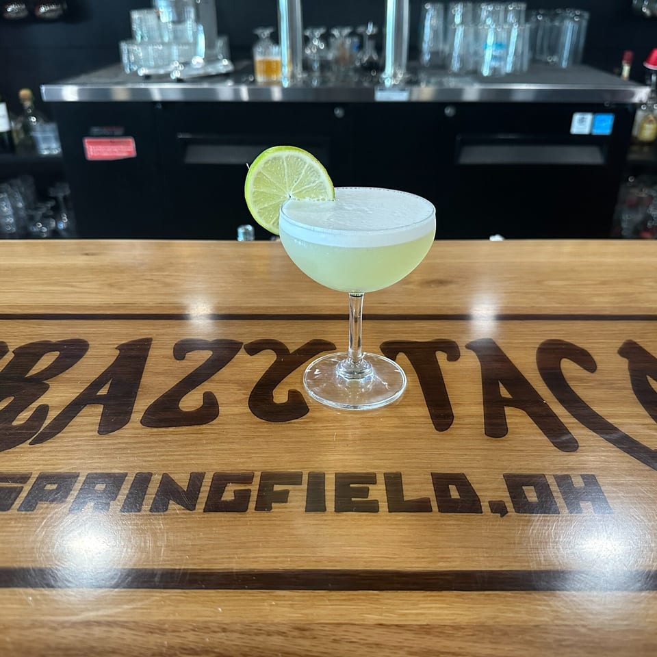 A Krazy Taco 937 margarita with a lime wedge sits on a wooden bar with the restaurant's logo.