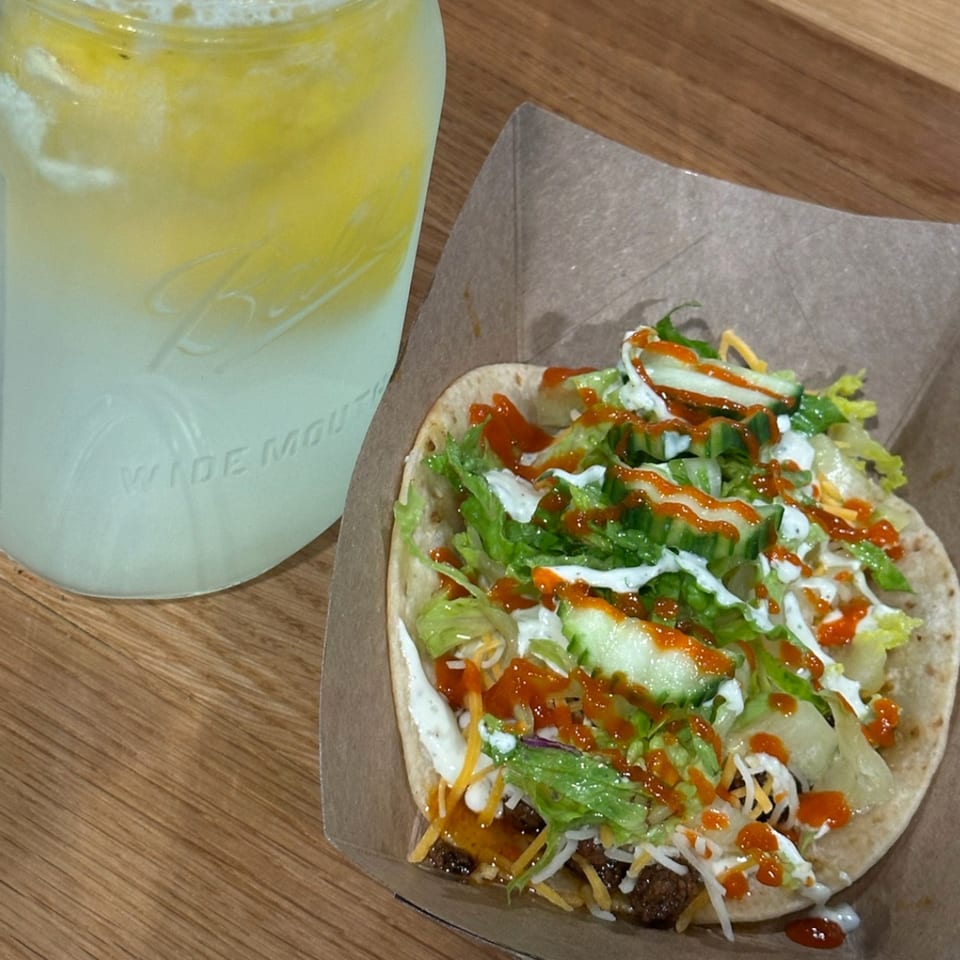A Krazy Taco 937 taco with lettuce, cheese, sauce, and cucumbers next to a lemonade.